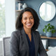 A friendly, diverse woman smiling confidently in a modern, bright office setting with soft blue-toned decor.